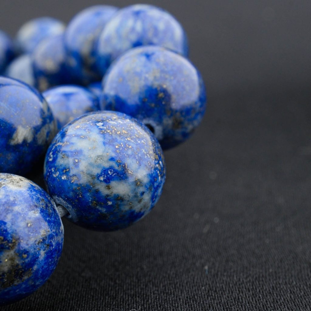 A horizontal row of square-cut lapis lazuli beads in deep blue, showing natural golden pyrite specks and light grey marbling against a white background.
