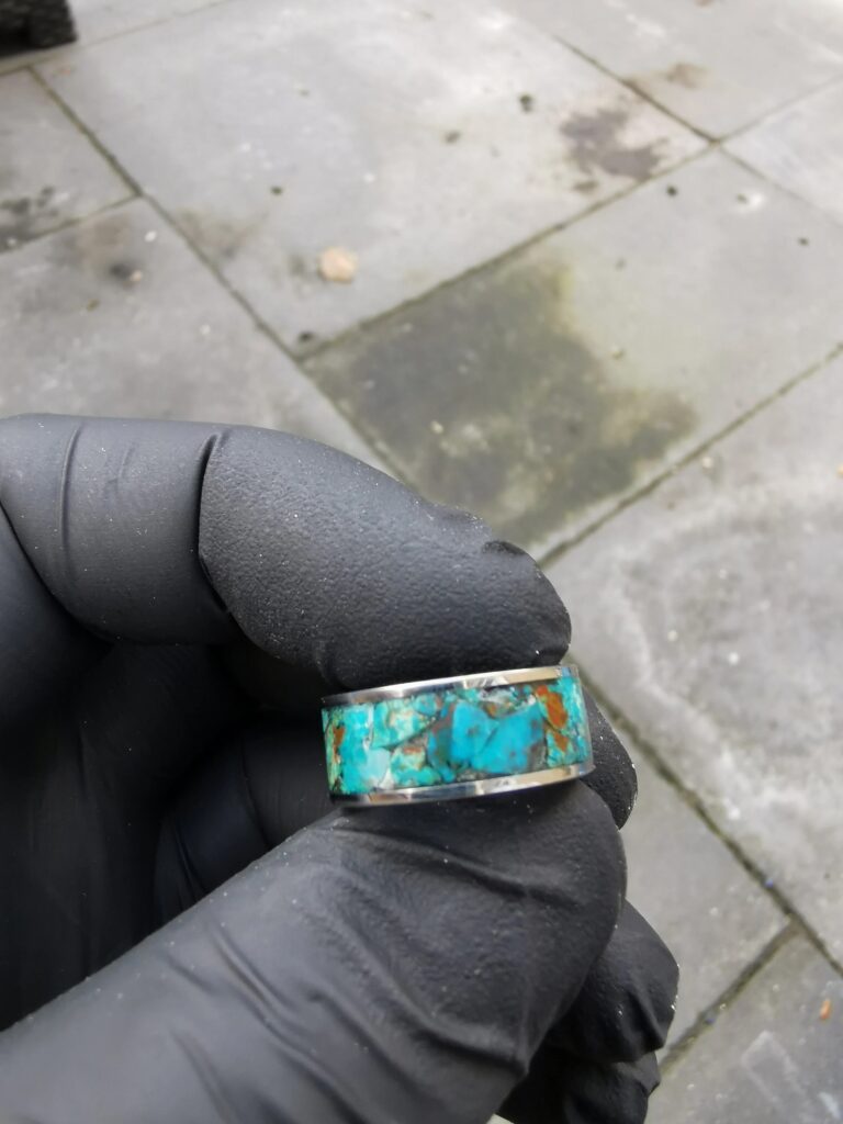 A close-up of a person wearing a black glove, holding a silver band ring inlaid with fragments of blue and brown chrysocolla.